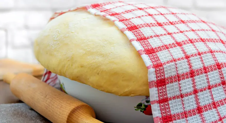 Puszyste ciasto drożdżowe rośnie w misce, przykryte kratkowaną ściereczką. Obok wałek do ciasta i rozsypana mąka.
