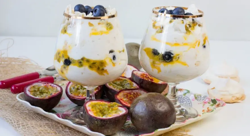 Dwa puchary z Eton Mess, przełożone kremem, marakują i borówkami, ozdobione bezą. Obok owoce marakui i bezy.