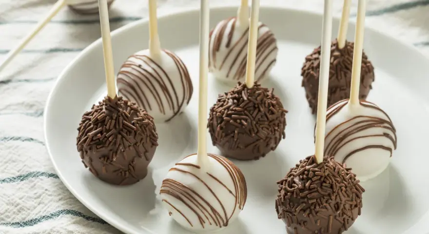 Słodkie cake pops na patyczkach, niektóre w czekoladzie z posypką, inne w białej polewie z czekoladowymi paskami.