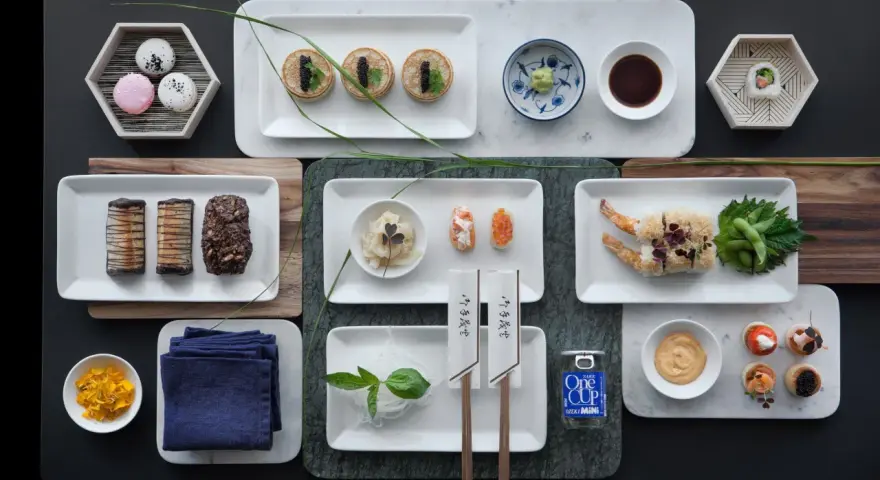 Elegancka aranżacja stołu z różnorodnymi przekąskami, w tym mini naleśnikami z kawiorem, sushi, krewetkami w tempurze i makaronikami. Idealne na wieczór, gdy masz ochotę na coś pysznego do sushi.