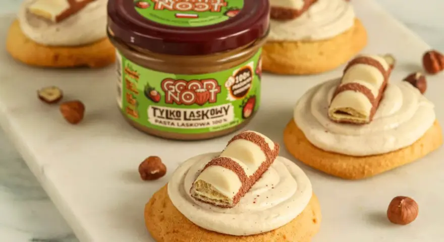 Pyszne ciasteczka inspirowane przepisem na crumbl cookies, z kremem i kawałkami czekolady. Słoik pasty z orzechów laskowych w tle.
