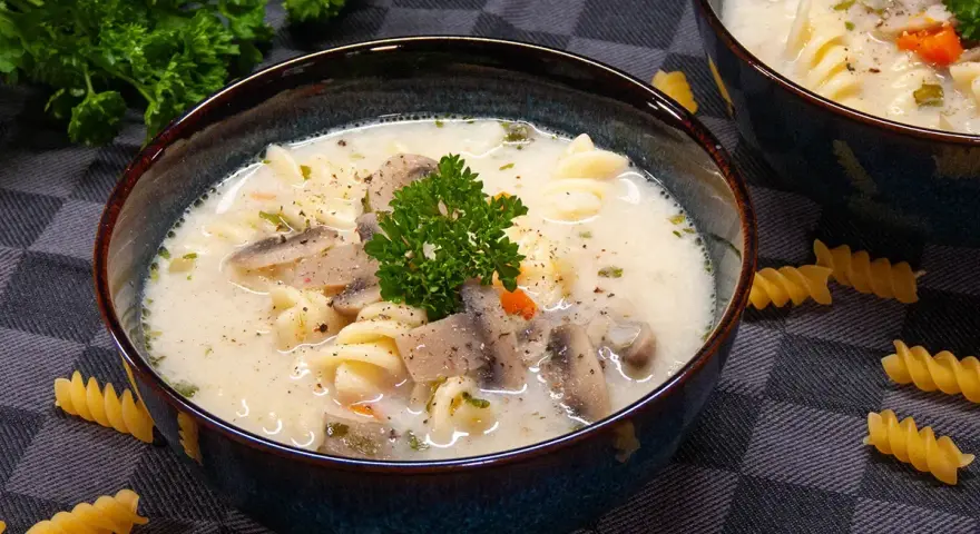 Aksamitna zupa pieczarkowa z makaronem, udekorowana natką pietruszki. Na stole leżą spiralne fusilli, zachęcając do spróbowania.