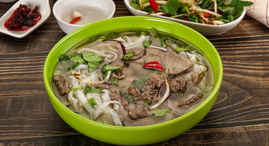 A miseczka aromatycznej zupy pho z wołowiną, makaronem ryżowym, świeżą kolendrą i chili. Obok sosy i dodatki.