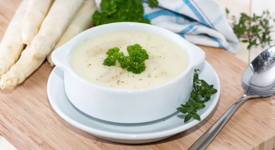 Aksamitna zupa szparagowa, udekorowana natką pietruszki, obok świeżych szparagów i łyżki.