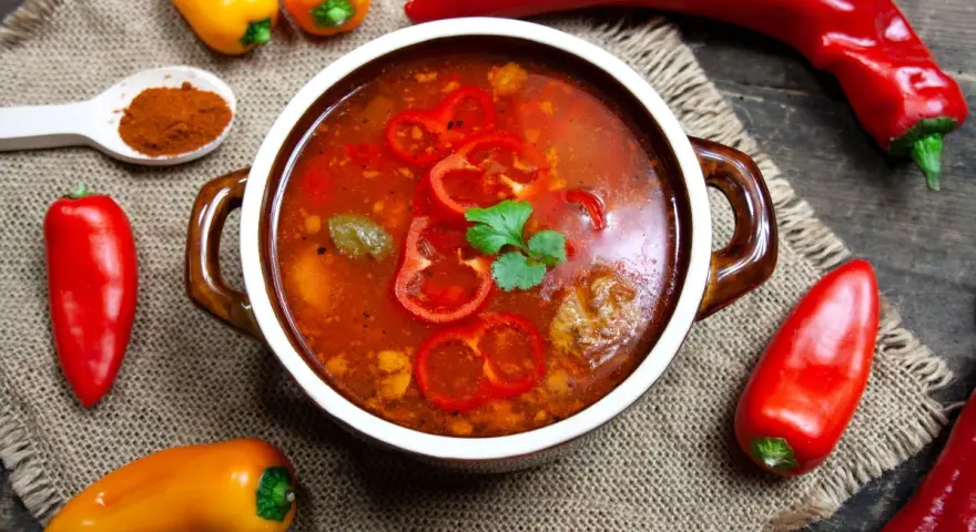Aromaticzna zupa gulaszowa w misce, otoczona papryczkami chili i słodkimi paprykami. Na łyżce mielona papryka.