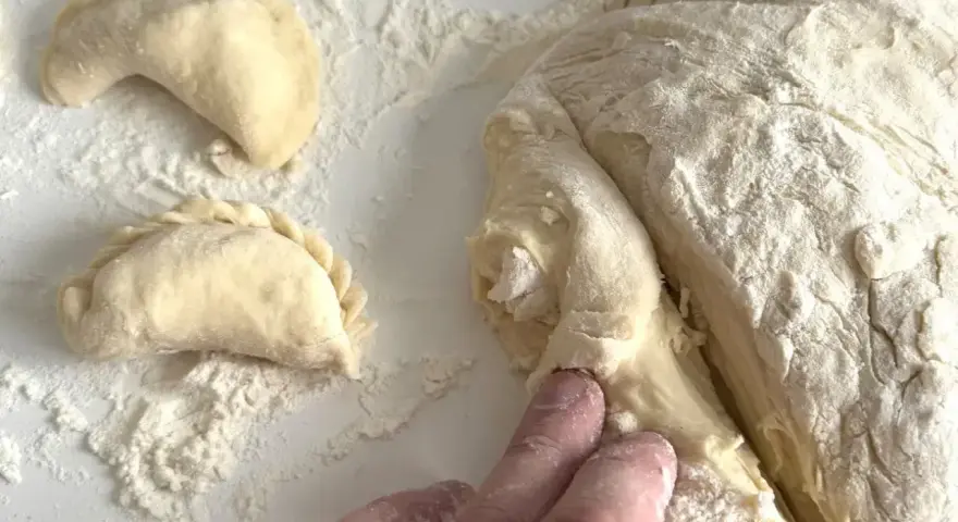 Ręce formują ciasto na pierogi, tworząc półksiężycowate kształty. Mąka rozsypana na blacie.