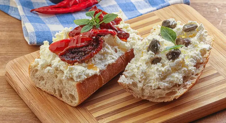 Dwa kawałki bagietki z twarogiem, suszonymi pomidorami i kaparami. Kapary dodają wyrazistego smaku.