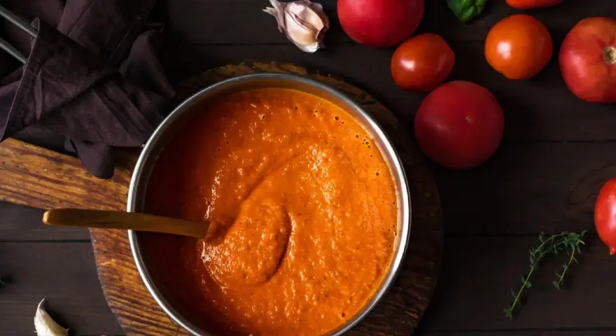 Gęsty, pomidorowy sos w garnku, obok świeże pomidory, czosnek i bazylia. Idealny przepis na to, jak zagęścić sos pomidorowy.