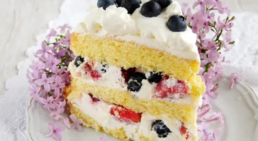 Pyszny tort z bitą śmietaną i mascarpone, przełożony owocami leśnymi, ozdobiony jagodami i kwitnącym bzem.
