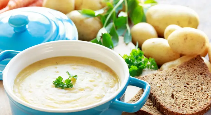 Aksamitna zupa ziemniaczana w niebieskiej misce, udekorowana natką pietruszki. Obok surowe ziemniaki, marchewki i kromki ciemnego chleba.