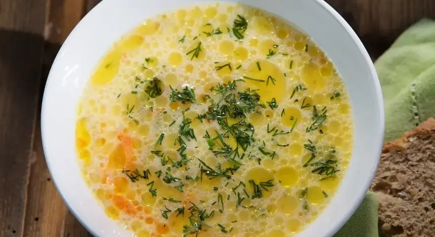 Aromaticzna zupa ogórkowa, pełna koperku i pływających kuleczek tłuszczu, czeka na talerzu obok kromki chleba.