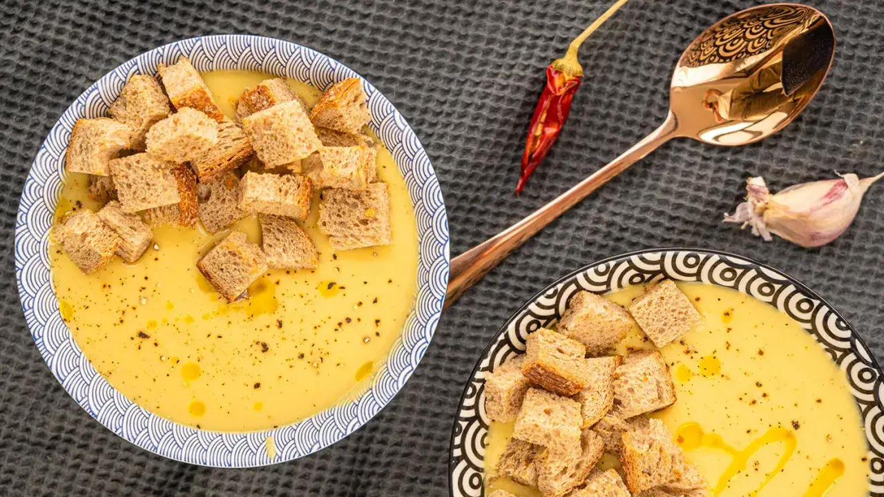 Dwie miseczki kremowej zupy z ciecierzycy, posypanej grzankami. Obok leży suszona papryczka chili, ząbek czosnku i elegancka łyżka.