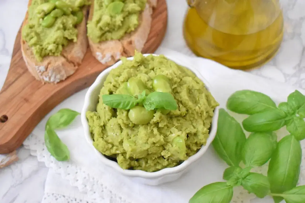 Pyszne pesto z bobu w miseczce, ozdobione listkami bazylii i fasolkami. Obok kromki chleba z pastą i butelka oliwy.