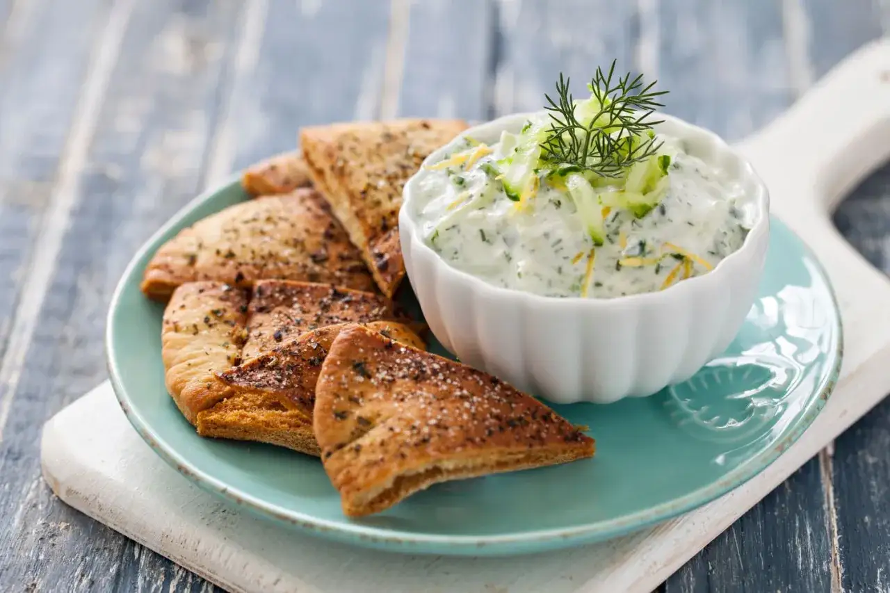 Chrupiące kawałki pity z przyprawami i miseczka kremowego sosu tzatziki, udekorowana świeżym koperkiem i ogórkiem.