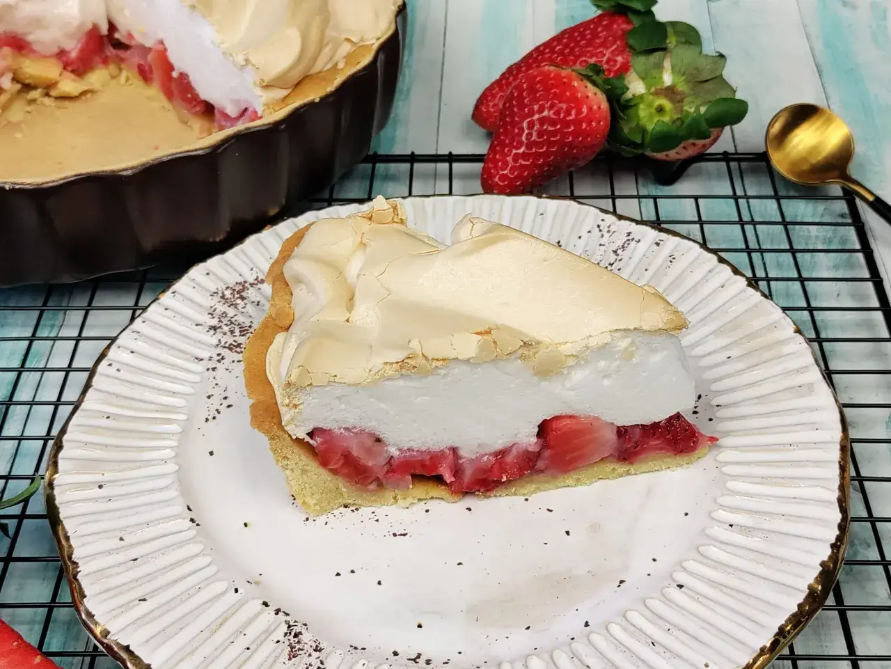 Pyszna tarta z truskawkami i bezą, z kawałkiem na talerzu. Obok świeże truskawki i złota łyżeczka.