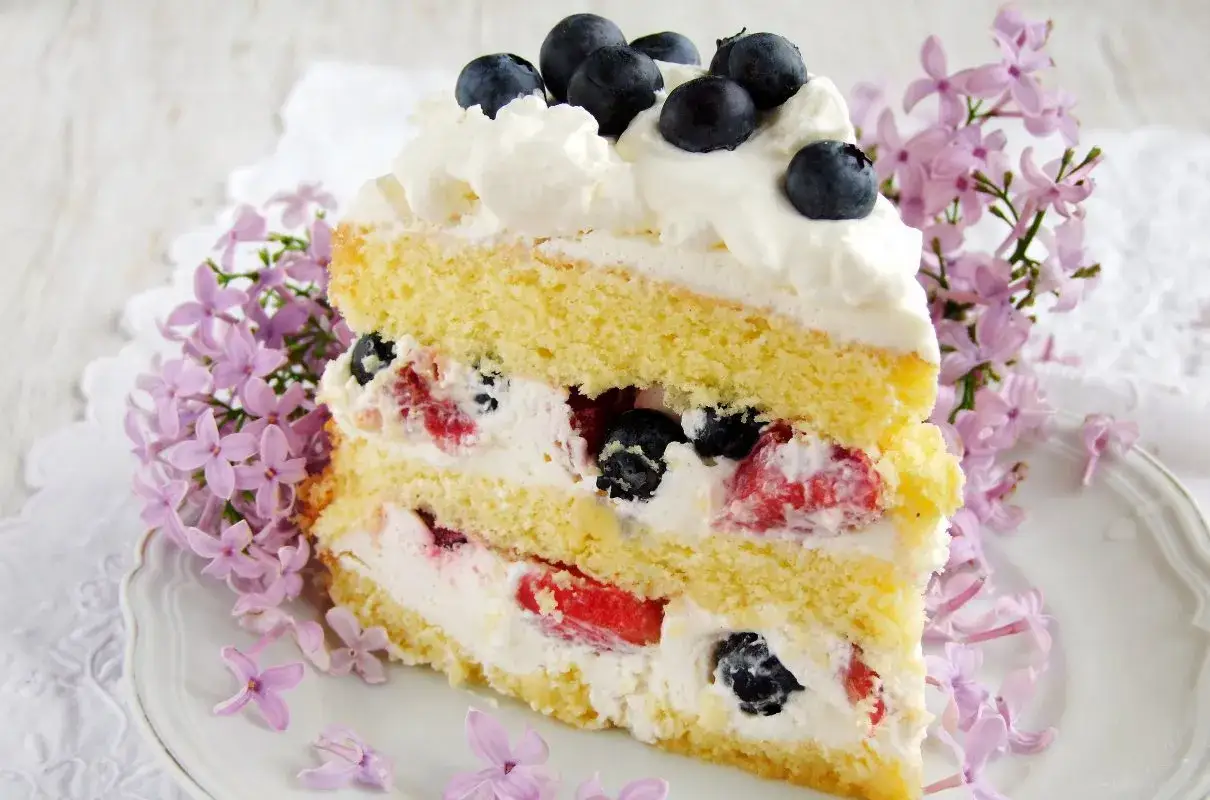 Pyszny tort z bitą śmietaną i mascarpone, przełożony owocami leśnymi, ozdobiony jagodami i kwitnącym bzem.