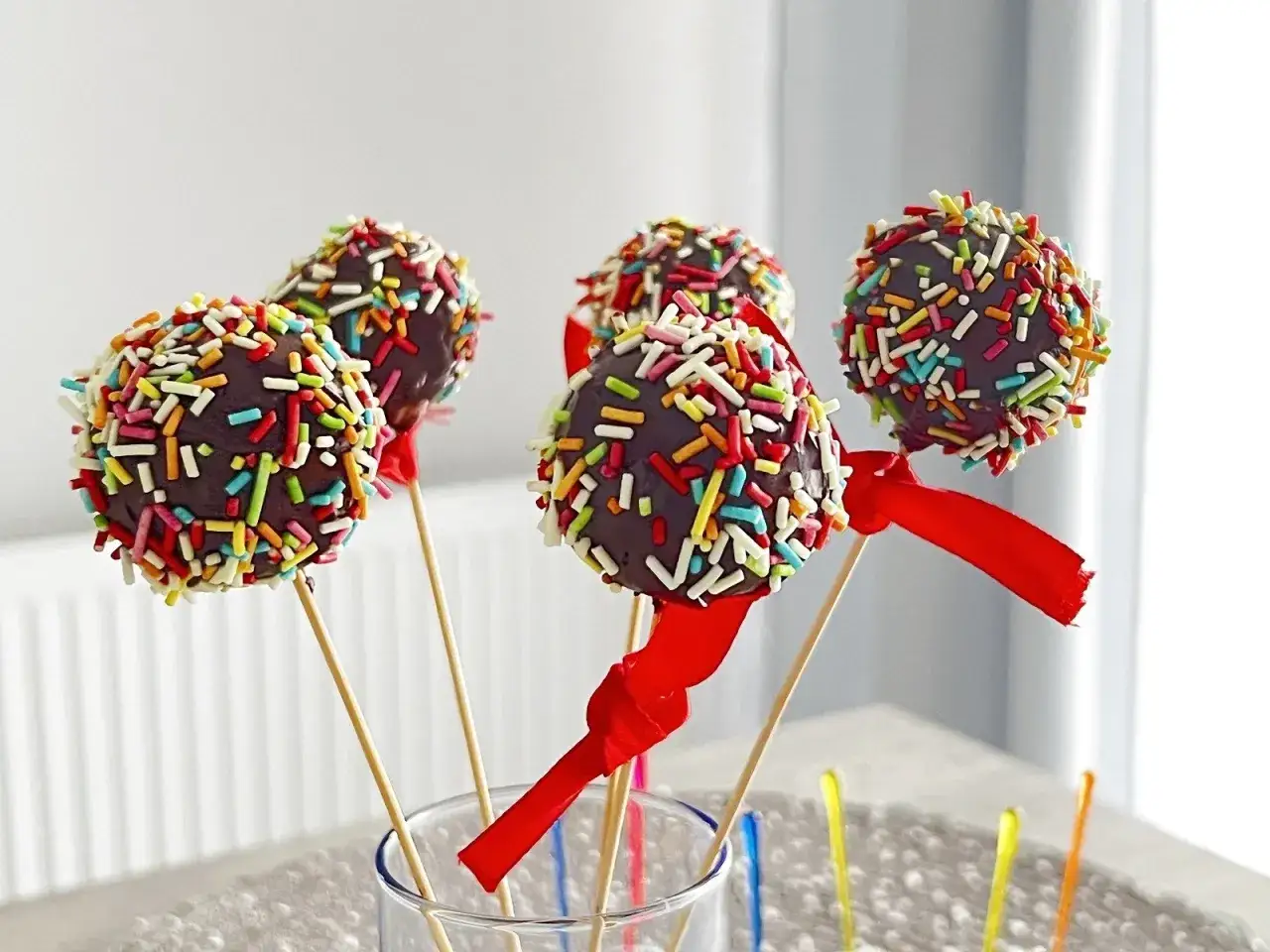 Kolorowe cake pops w czekoladzie, ozdobione kolorową posypką i czerwonymi wstążeczkami, stoją w szklance.