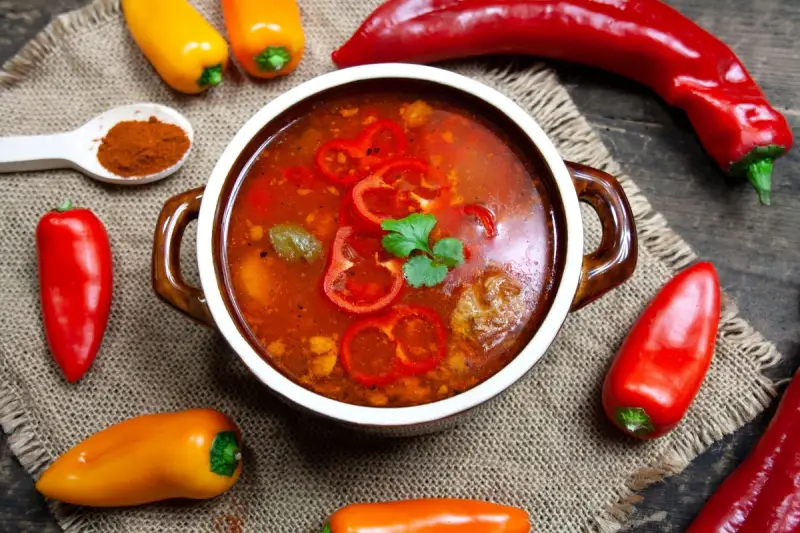 Aromaticzna zupa gulaszowa w misce, otoczona papryczkami chili i słodkimi paprykami. Na łyżce mielona papryka.